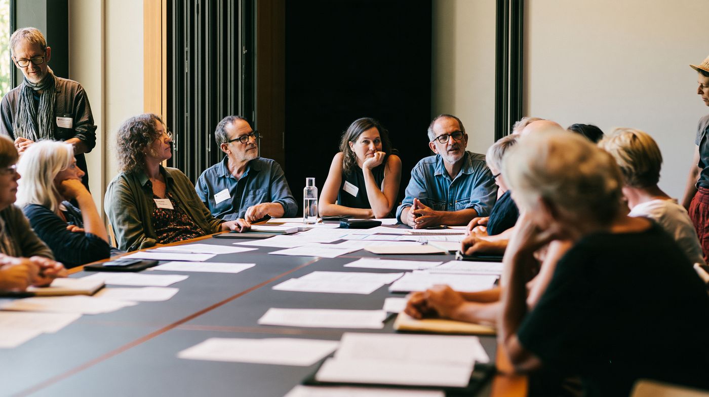 A group of people seated around a long table with papers, listening to one another